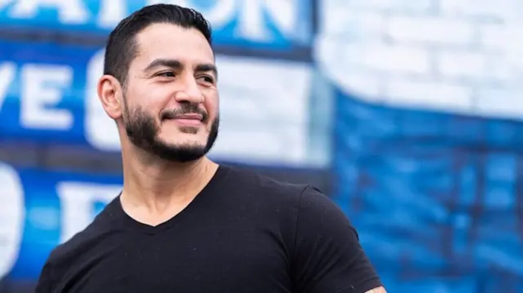 Abdul El-Sayed sonríe de perfil, vestido con una camiseta negra, frente a un fondo azul.