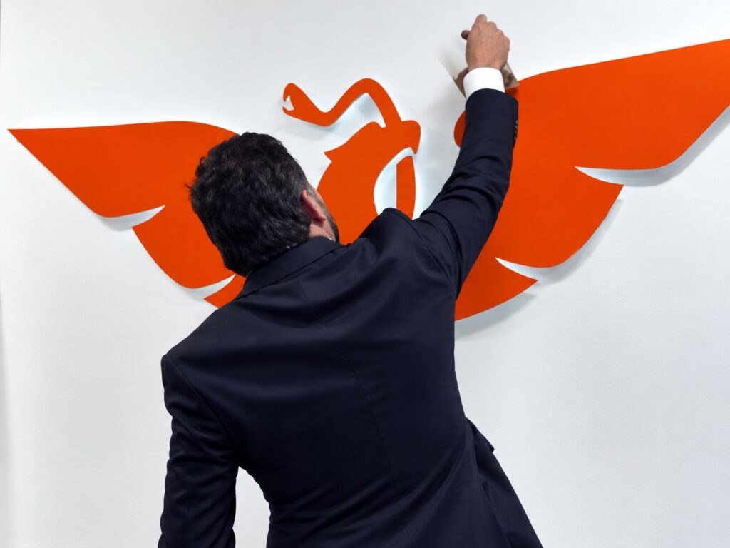 Persona de espaldas con el brazo en alto frente al logotipo naranja de Movimiento Ciudadano en una pared blanca
