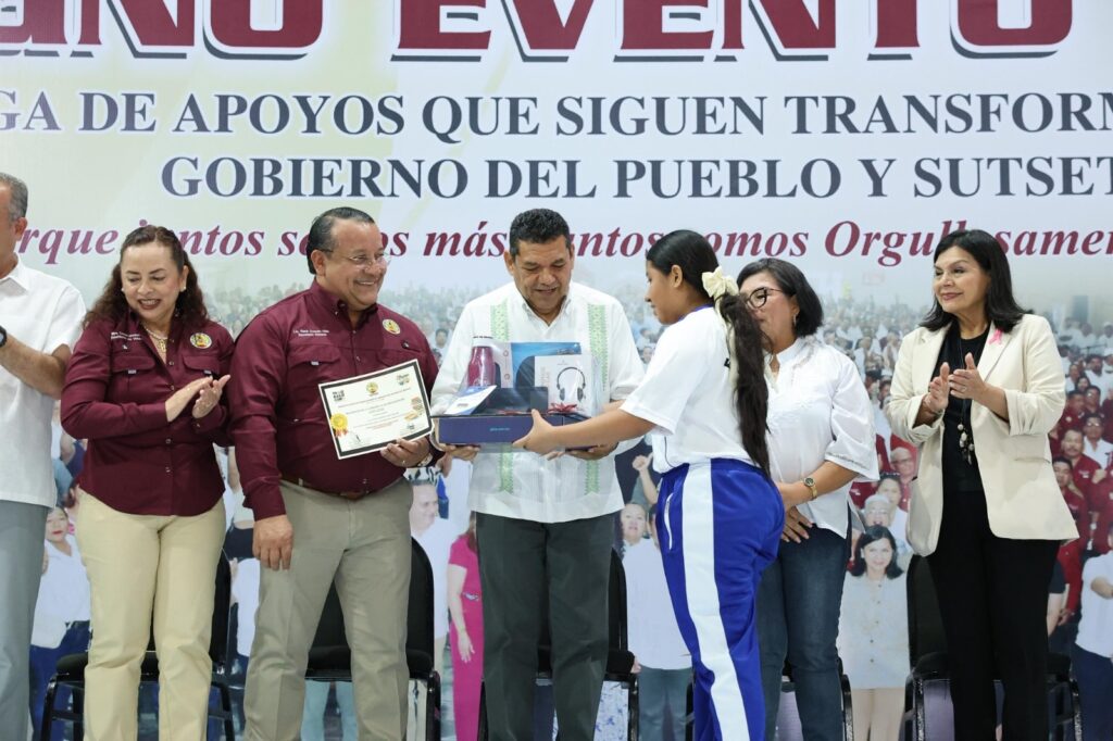 Alumna en uniforme recibe un paquete con laptop, audífonos y botella en el escenario del Magno Evento 2026, mientras asistentes en el estrado aplauden