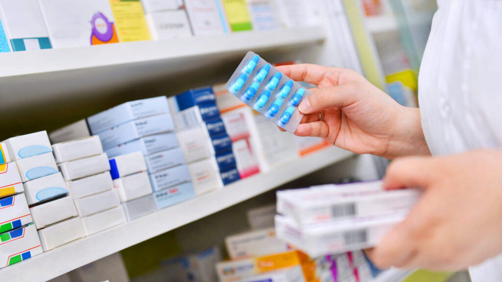 Mano sostiene un blíster de cápsulas azules frente a un anaquel con cajas de medicamentos en una farmacia.