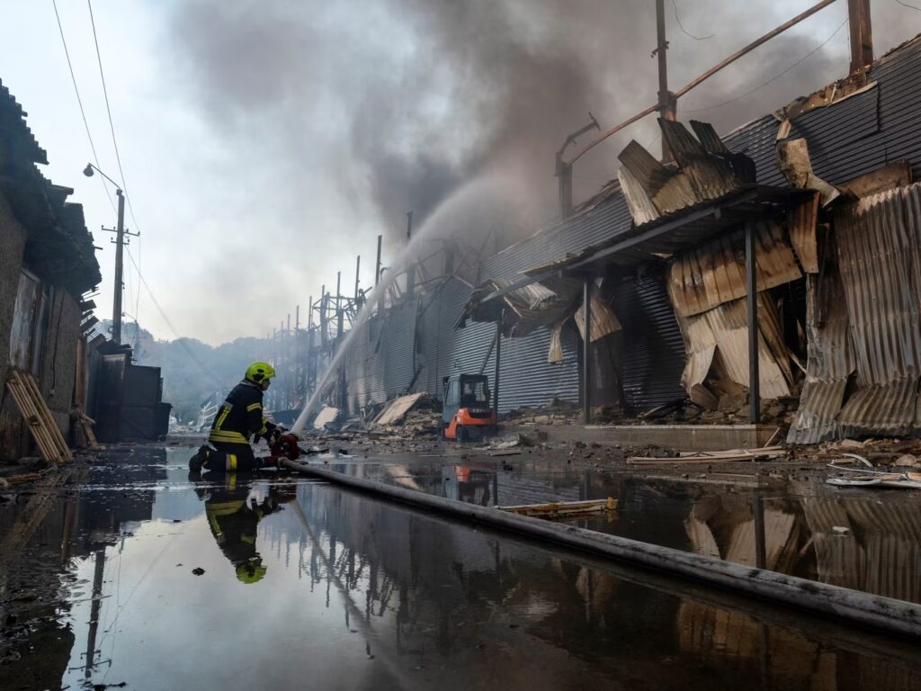 Bombero ucraniano combate un incendio entre estructuras metálicas destruidas, escombros y agua acumulada.