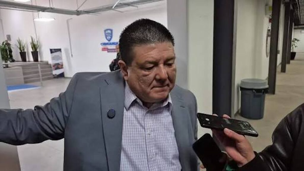 Hombre con saco gris hablando con periodistas que acercan grabadoras y teléfonos móviles para registrar sus declaraciones.
