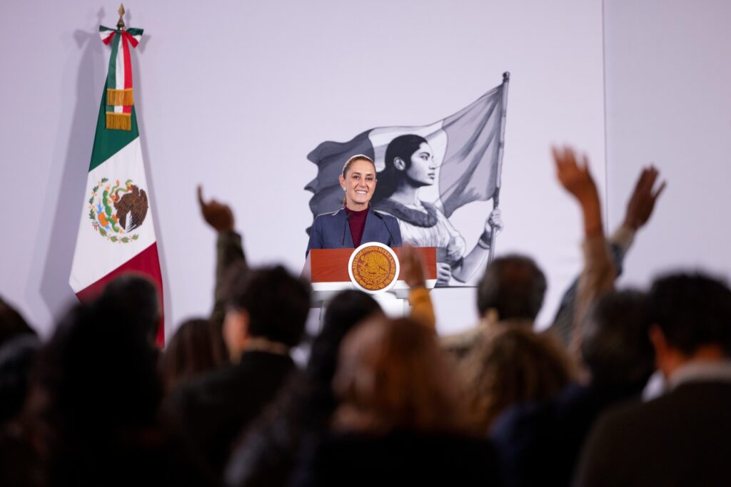 Claudia Sheinbaum responde preguntas de periodistas durante su conferencia matutina, frente a la bandera mexicana y el podio presidencial.