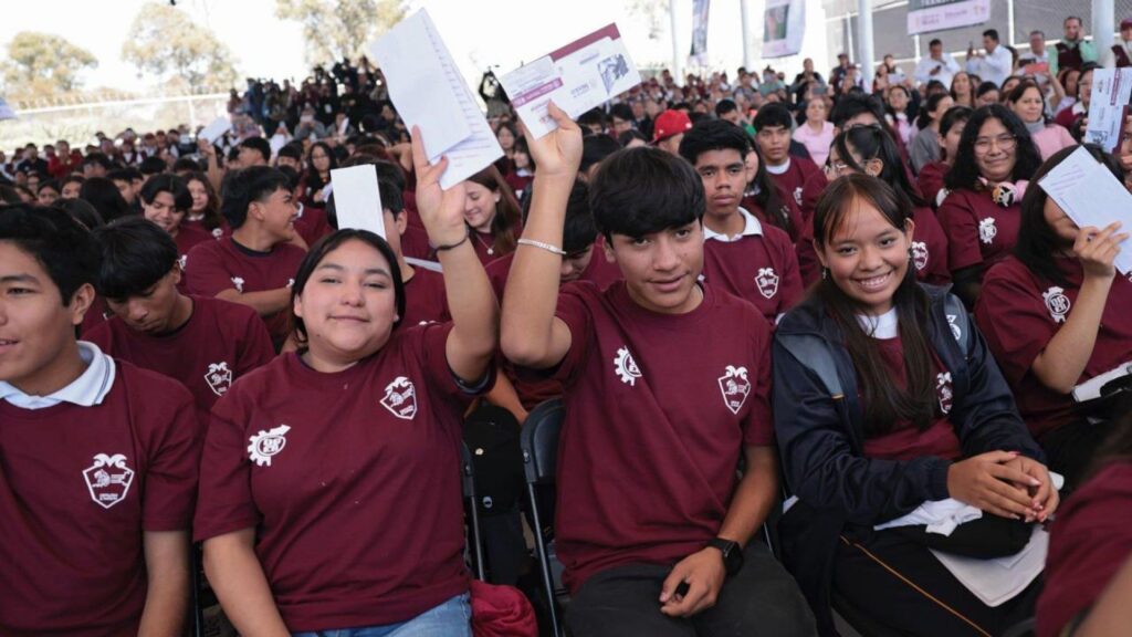 Estudiantes de bachillerato con playera guinda levantan sobres con sus tarjetas durante un evento de becas