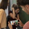 Adolescente graba con su celular a una compañera que se cubre el rostro contra una pared mientras otra la observa