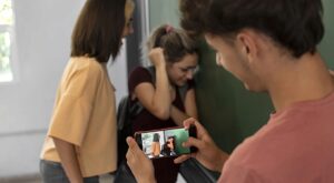 Adolescente graba con su celular a una compañera que se cubre el rostro contra una pared mientras otra la observa