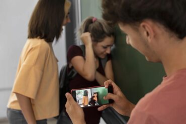 Adolescente graba con su celular a una compañera que se cubre el rostro contra una pared mientras otra la observa