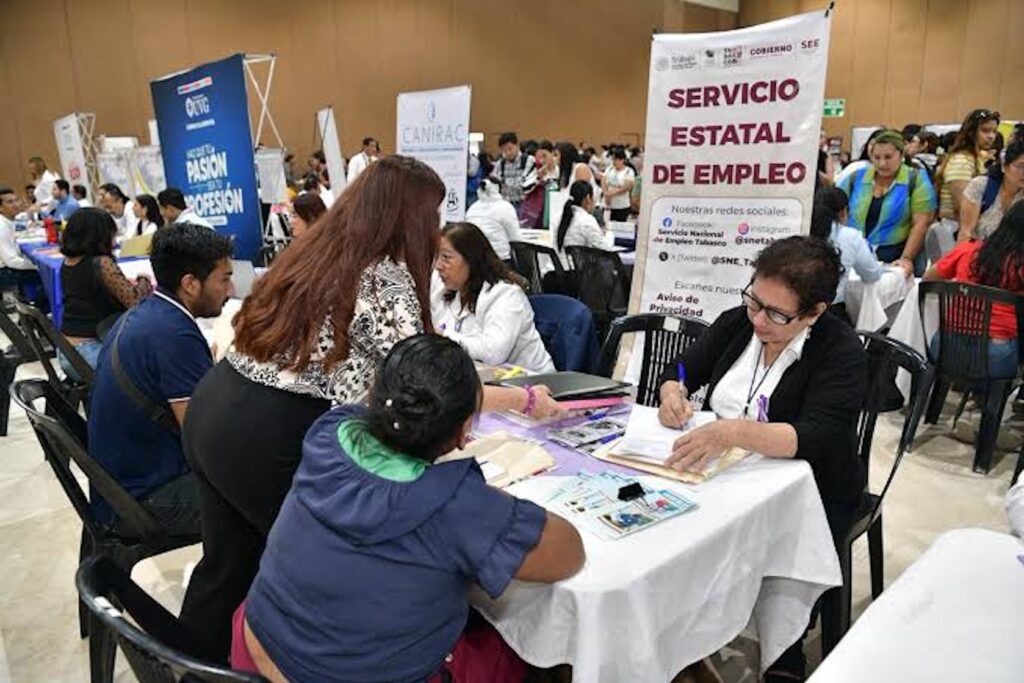 Personas solicitantes son atendidas en mesas del Servicio Estatal de Empleo durante una feria de empleo en un salón con módulos de empresas
