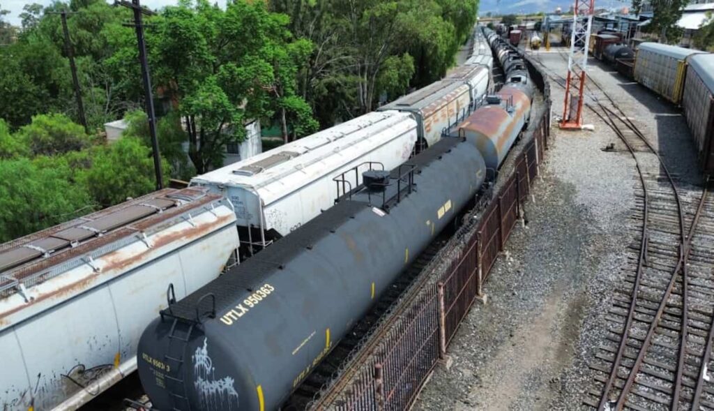 Vista aérea de ferrotanques y tolvas en un patio ferroviario, con vías y una torre al fondo.