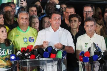Flávio Bolsonaro lee un documento ante micrófonos de medios, rodeado de aliados con camisetas de su campaña, tras perder la segunda vuelta ante Lula