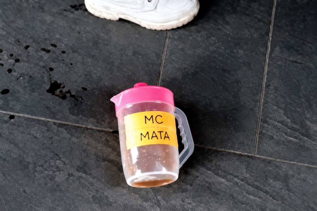 Jarra de plástico con tapa rosa y etiqueta “MC MATA” tirada en el piso, con líquido oscuro derramado