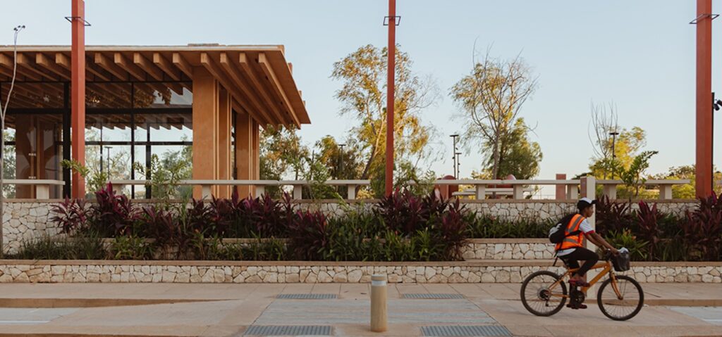 Ciclista con chaleco naranja frente a un pabellón de madera y jardineras de piedra en un paseo peatonal.
