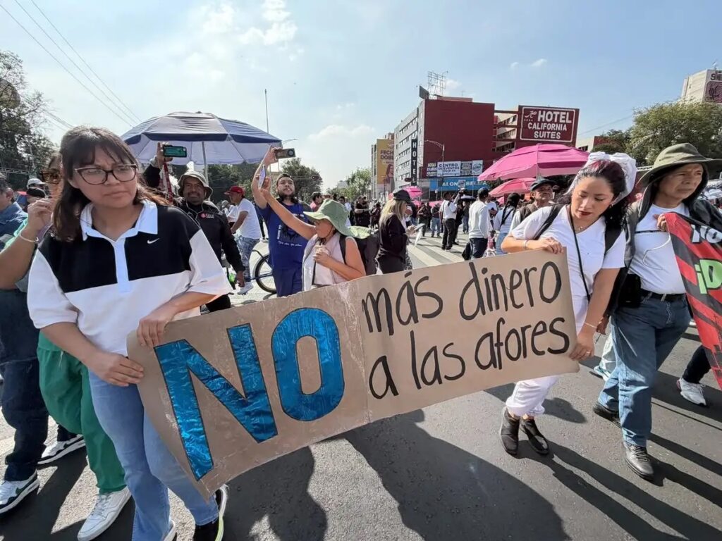 Dos mujeres con ropa clara sostienen una pancarta de cartón con la leyenda «NO más dinero a las afores» durante la marcha de trabajadores del IMSS.