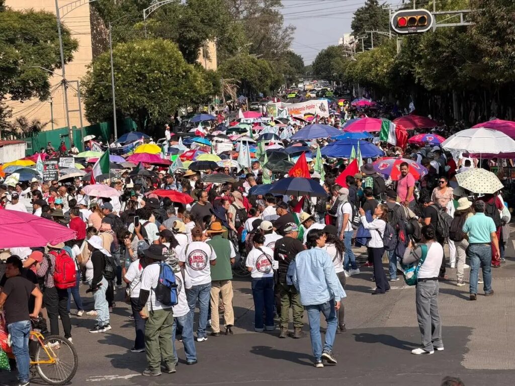 Contingente numeroso con paraguas de colores, banderas y mantas avanza por una calle arbolada; algunos participantes visten playeras con el logotipo del IMSS.