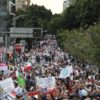 Multitud de trabajadores del IMSS con batas blancas avanza por una avenida arbolada durante la marcha nacional, con una bandera del instituto y carteles contra el dirigente sindical.