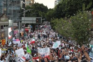 Multitud de trabajadores del IMSS con batas blancas avanza por una avenida arbolada durante la marcha nacional, con una bandera del instituto y carteles contra el dirigente sindical.