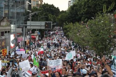 Multitud de trabajadores del IMSS con batas blancas avanza por una avenida arbolada durante la marcha nacional, con una bandera del instituto y carteles contra el dirigente sindical.