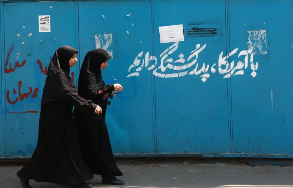 Dos mujeres con chador negro caminan frente a un muro azul con grafiti en persa en una calle de Irán