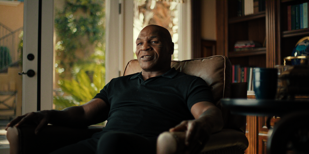 "A man sitting in a cozy armchair in a warmly lit home, surrounded by bookshelves and large windows with sunlight and plants visible outside, a coffee table with a mug in the foreground."