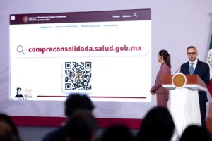 Pantalla de la mañanera con la Plataforma de compras consolidadas del sector Salud, su dirección web compraconsolidada.salud.gob.mx y un código QR, junto a dos funcionarios.