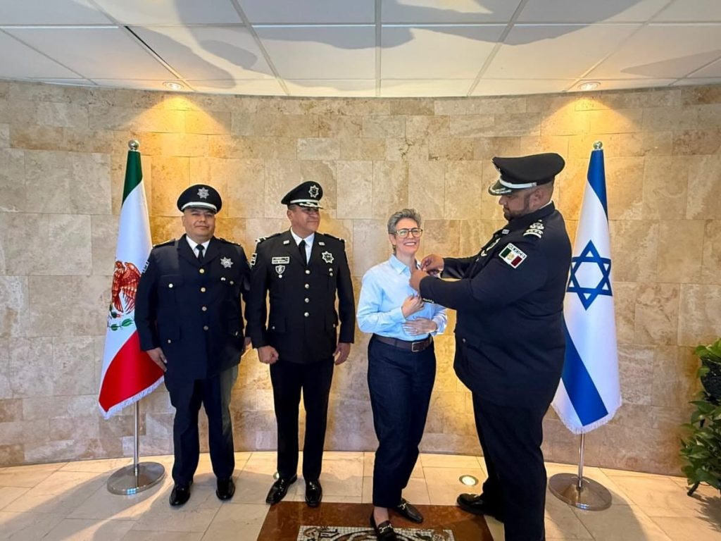 Integrantes uniformados de Policías Celestiales acompañan a una mujer durante la colocación de una insignia, frente a las banderas de México e Israel.