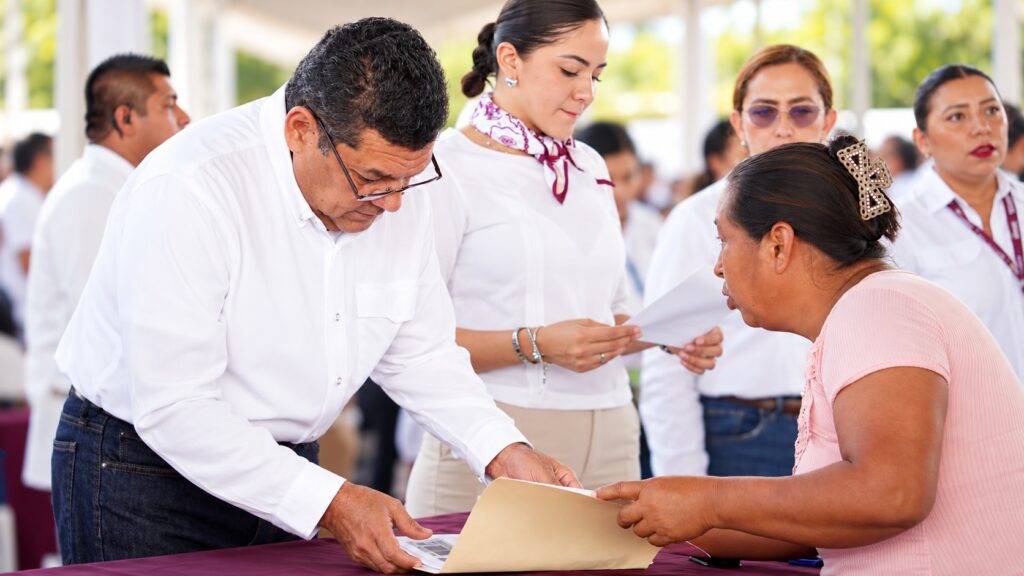 El gobernador Javier May, de camisa blanca y lentes, se inclina sobre una mesa para revisar documentos con una mujer sentada; al fondo, la titular de la Oficina de la Gubernatura, Sylvia Aylin Olvera Pérez, durante un evento al aire libre.