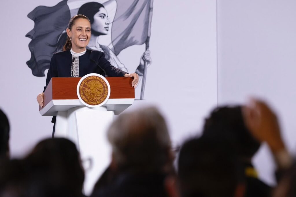 Claudia Sheinbaum sonríe en el podio con el escudo nacional frente a reporteros durante la conferencia matutina