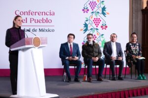 La presidenta Claudia Sheinbaum habla desde el podio durante la conferencia matutina, con cuatro asistentes sentados detrás