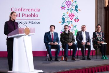 La presidenta Claudia Sheinbaum habla desde el podio durante la conferencia matutina, con cuatro asistentes sentados detrás