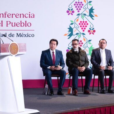 La presidenta Claudia Sheinbaum habla desde el podio durante la conferencia matutina, con cuatro asistentes sentados detrás