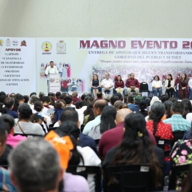 Orador en el podio del Magno Evento 2026 del SUTSET; el cartel del escenario enlista laptops, prótesis, licencias, gastos de titulación y lentes