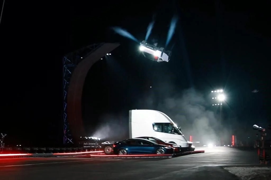 Tesla Roadster invertido en el aire con estelas de propulsores, junto a la rampa curva y sobre un camión Semi y dos autos