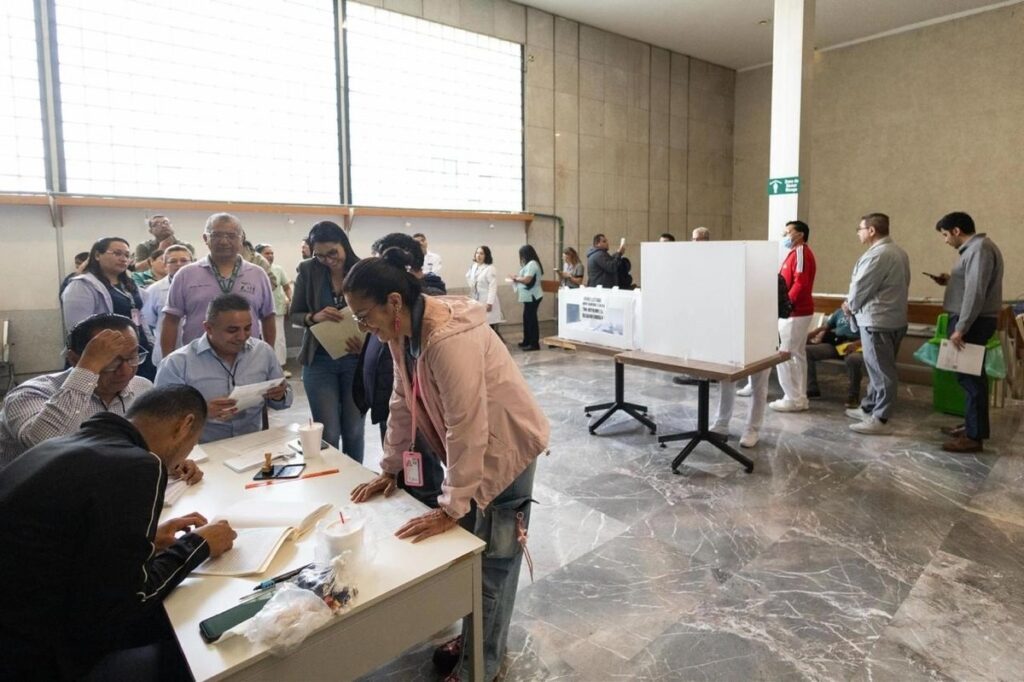 Trabajadores del IMSS hacen fila y firman en una mesa de registro junto a una mampara de votación blanca durante la consulta salarial.