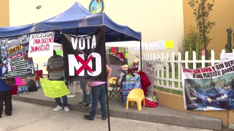 Plantón de trabajadores del IMSS bajo una carpa azul con una pancarta negra que dice «Vota NO» y letreros que exigen salarios dignos.