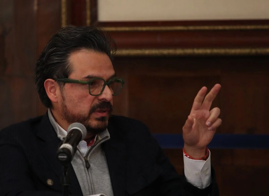 Zoé Robledo, director general del IMSS, habla frente a un micrófono y levanta la mano derecha